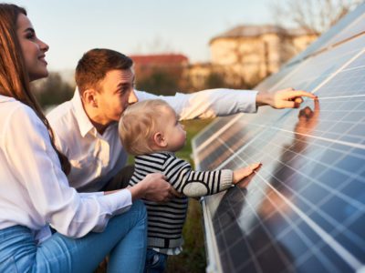 Side close-up shot of a young modern family with a little son getting acquainted with solar panel on a sunny day, green alternative energy concept
